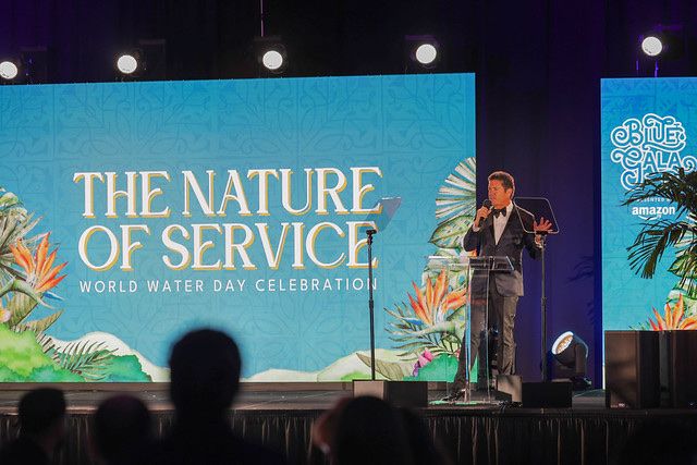 A speaker onstage before a screen reading The Nature of Service, World Water Day Celebration